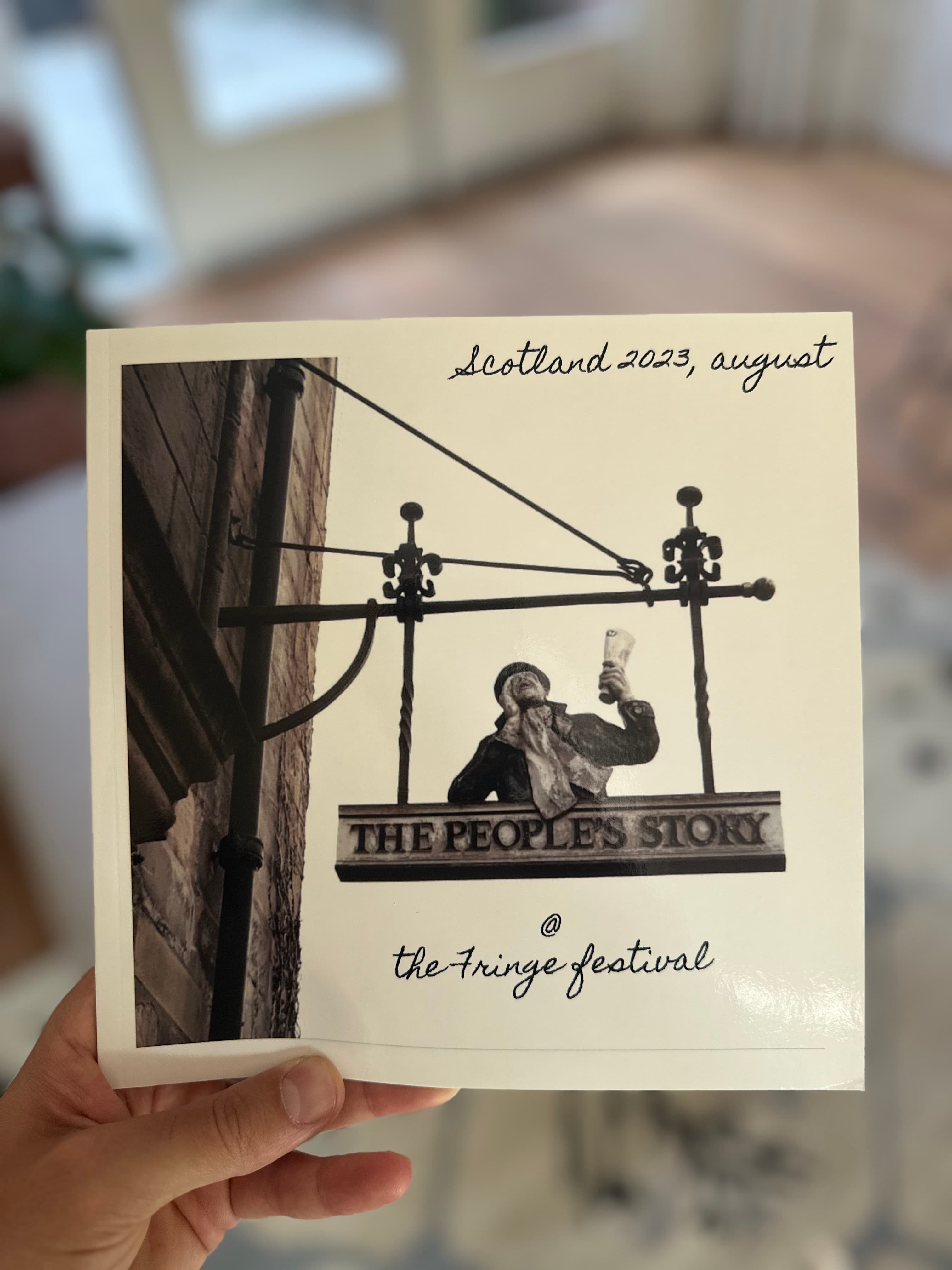 A hand holding a photobook whose black-and-white cover shows the hanging pub sign of The People's Story, handwritten "Scotland 2023, august" above it