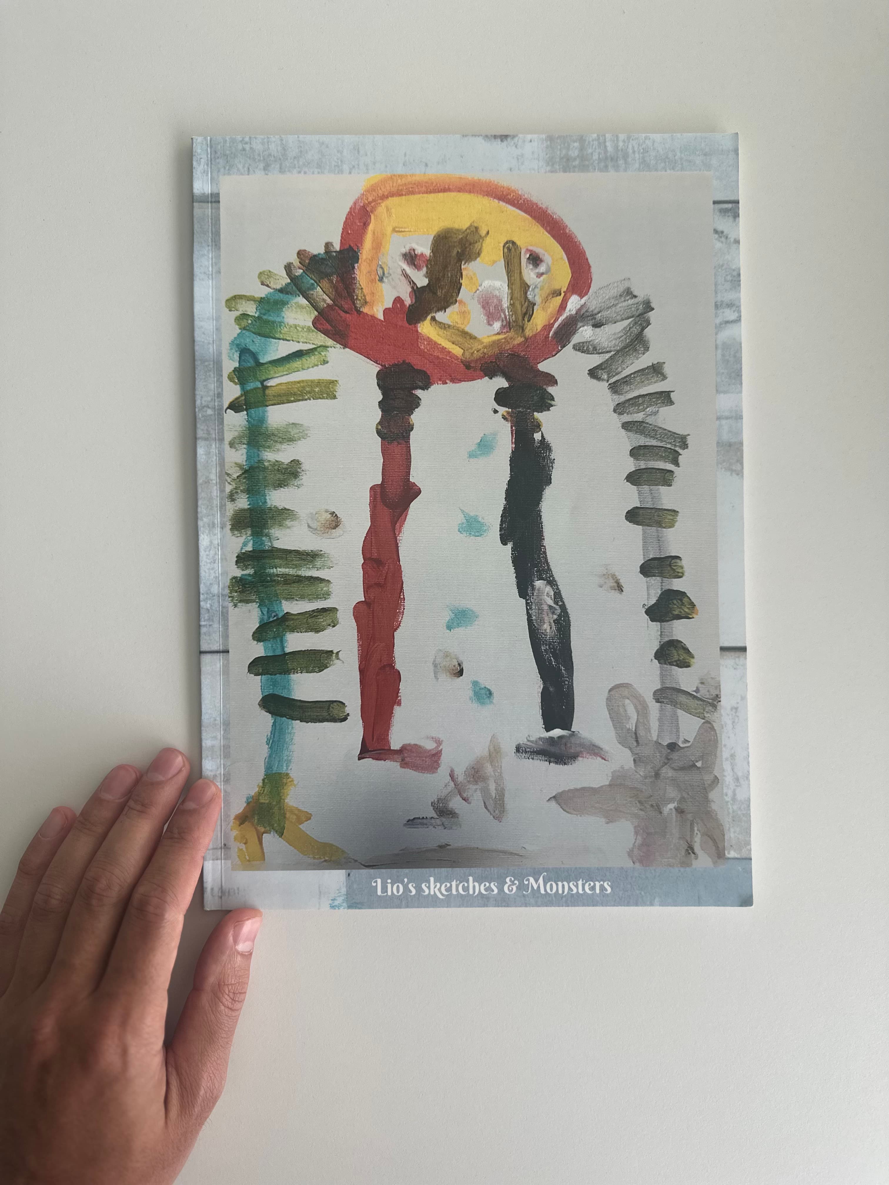A child's painting printed as a photobook cover, titled "Lio's sketches & Monsters", held up against a wall
